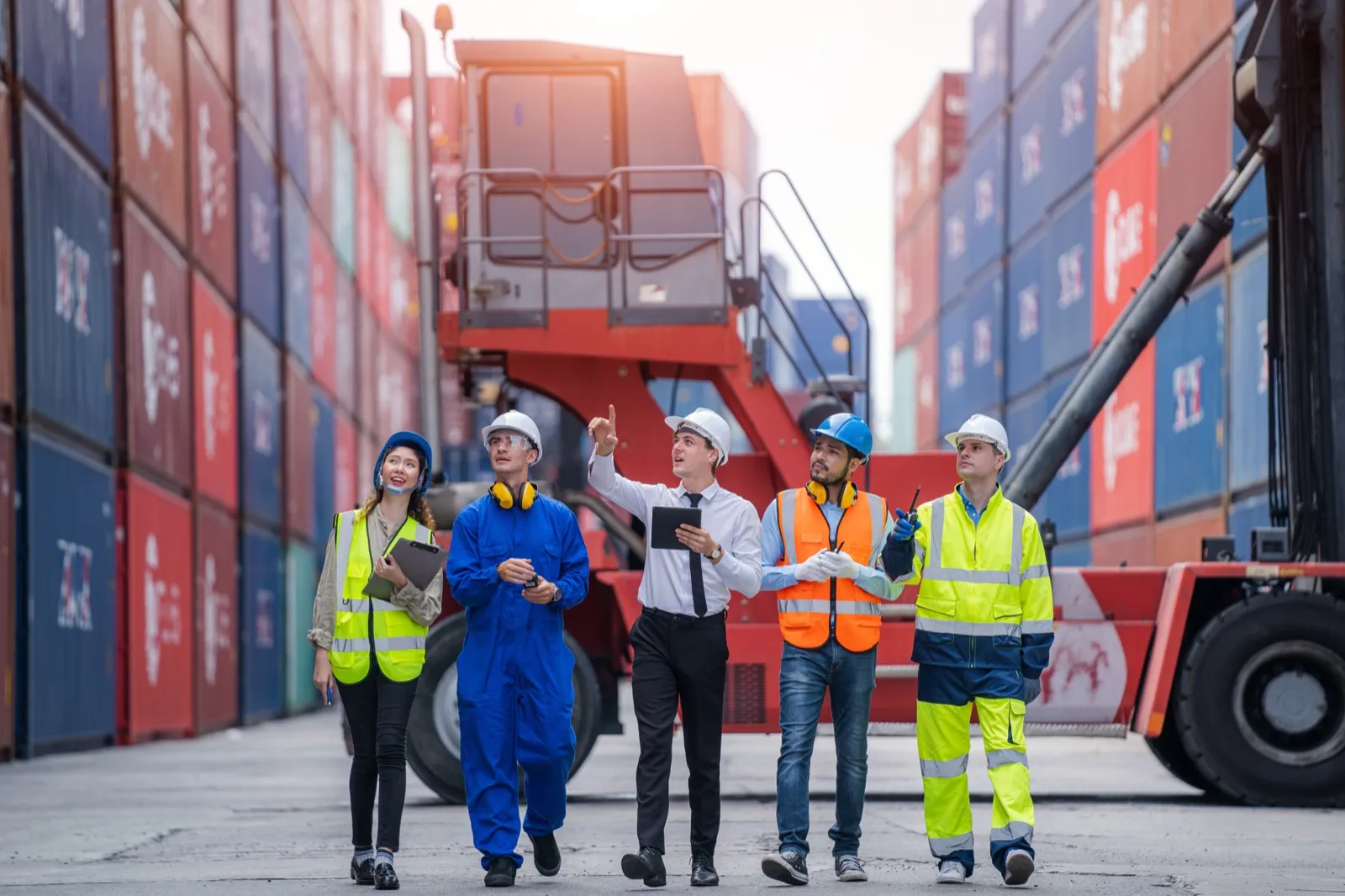Tre operatori con giubbotto ad alta visibilità camminano fra i container di un terminal.